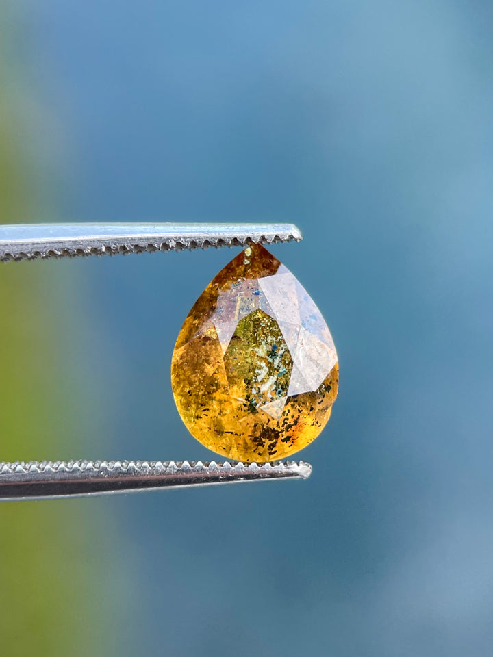 Close-up of a pear-shaped sapphire weighing 3.03 carats, displaying yellow, orange, and black inclusions, held with tweezers.