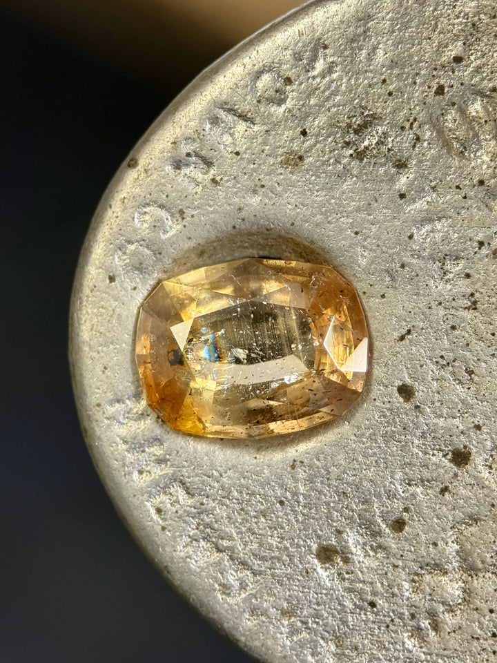  Faceted 4.02ct sapphire resting on a metallic surface, exhibiting warm golden peach and greenish-pink tones with natural inclusions and internal flashes.