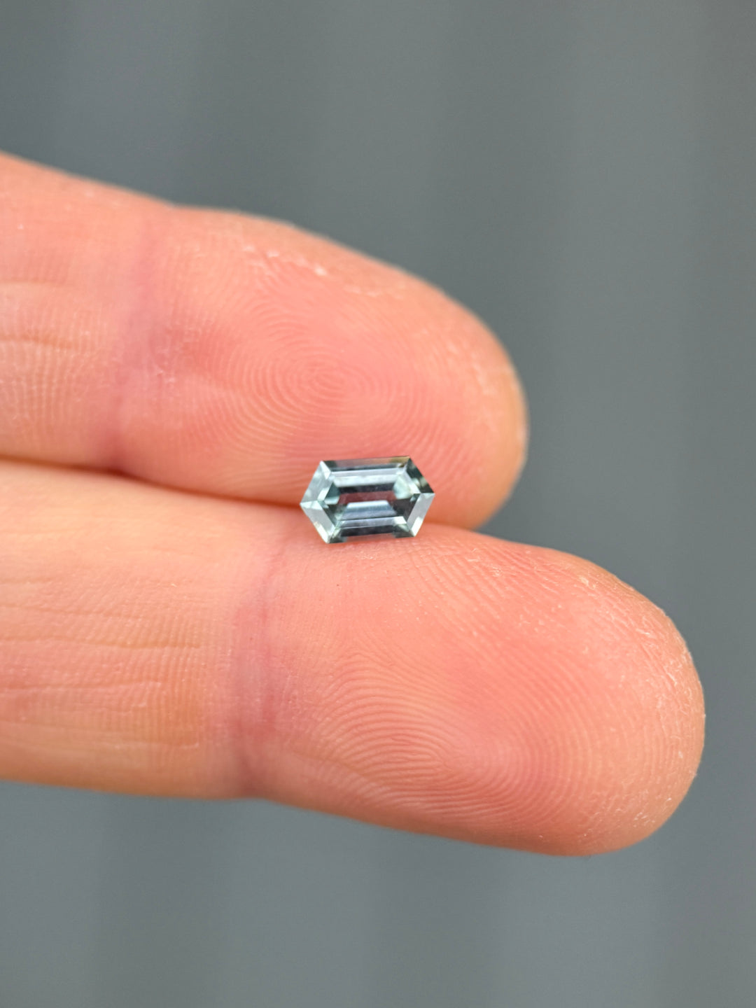 Macro photo of a color shift Montana sapphire resting on fingertips, revealing crisp elongated hexagon step cut facets and a transition from light seafoam to purple hues