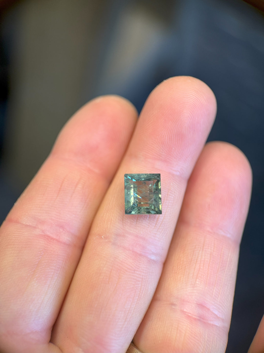 (Finger indoor light): Precision square tablet cut sapphire with green-blue hues on fingertip.