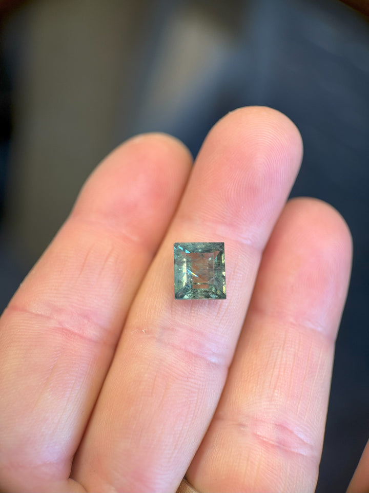 (Finger indoor light): Precision square tablet cut sapphire with green-blue hues on fingertip.