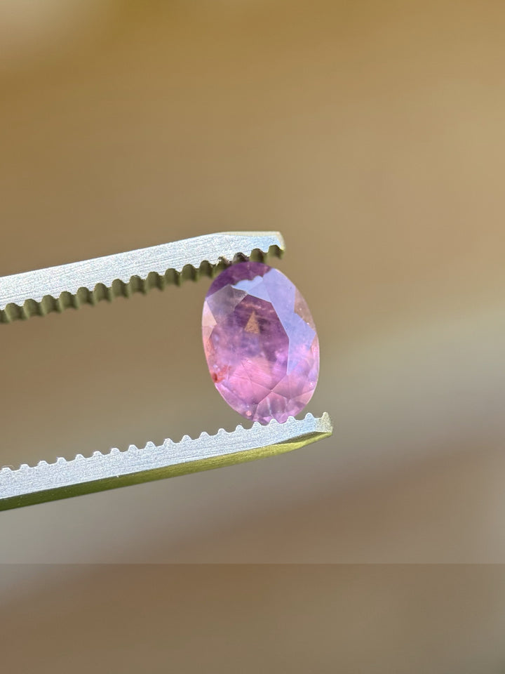 Unheated 1.37ct Umba sapphire held in tweezers with pink-purple hues visible through clean oval facets.