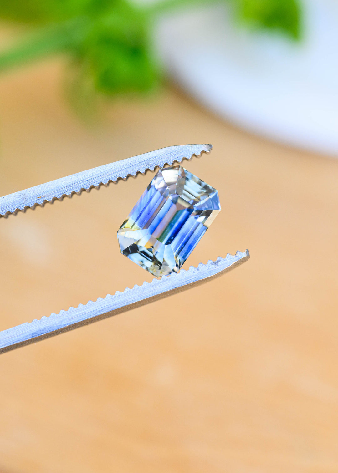 Unheated Sri Lankan parti sapphire held in jeweler’s tweezers, displaying an elongated emerald-cut shape with crisp clipped corners and alternating blue, yellow and near-colorless reflections.