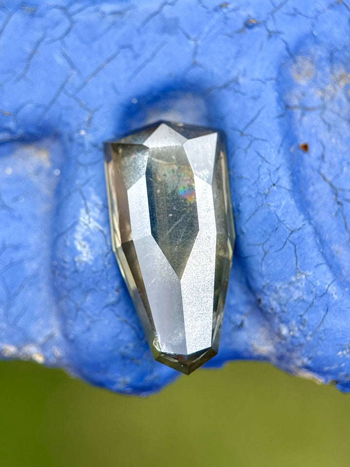 Color-shifting bluish-green sapphire with rainbow flash, held between Blue fingers to highlight brilliance.