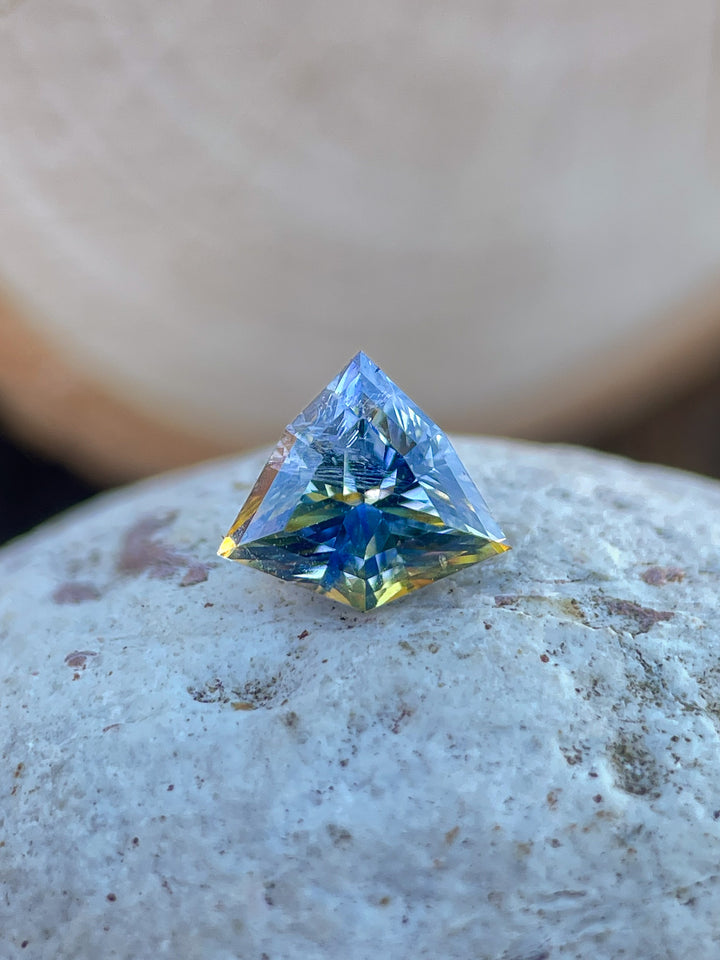 A beautifully faceted bi-color sapphire with vibrant yellow and cornflower blue tones, delicately held in tweezers against a neutral background.