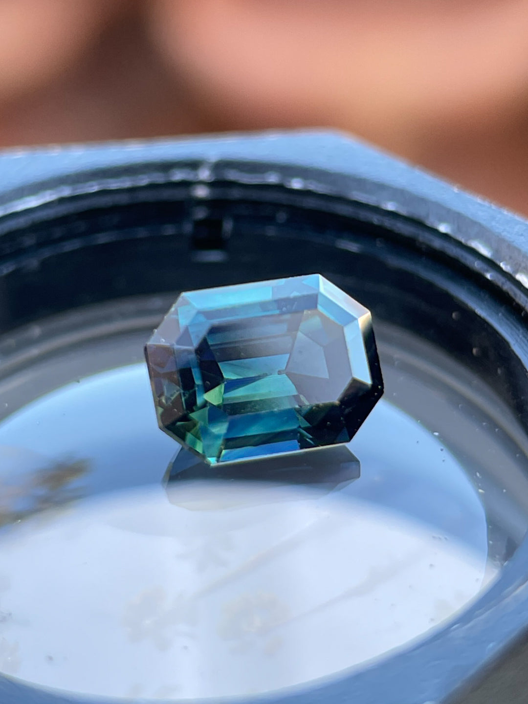 A beautiful bi-color green and blue step-cut sapphire resting on a reflective surface, emphasizing its clarity, origin from Gombe, Nigeria, and elegant cut.