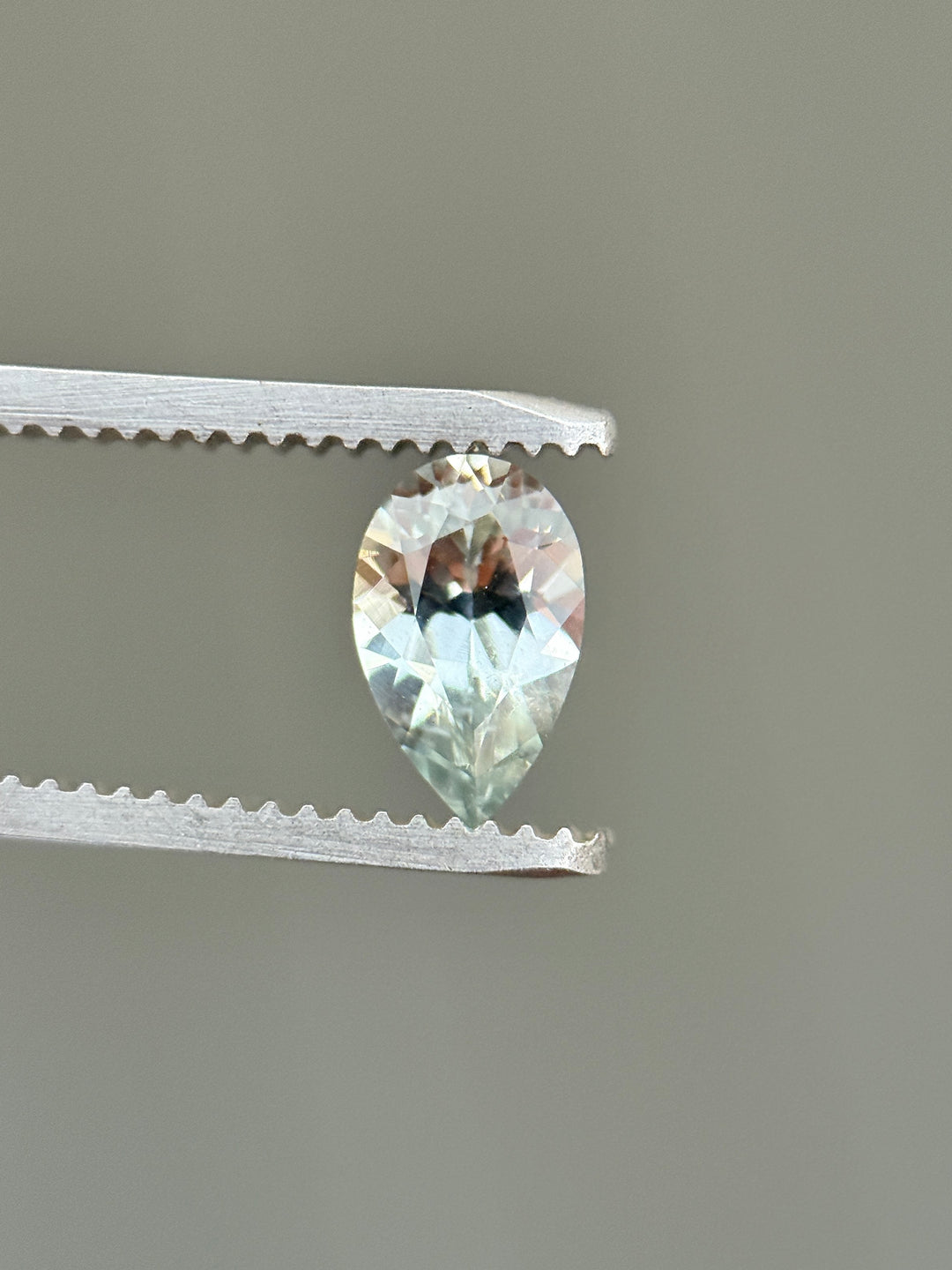 Side profile of a 1.13ct Montana sapphire held in gem tweezers, highlighting the precision pear mixed cut, pavilion depth, and subtle silver seafoam hue in an untreated natural sapphire.
