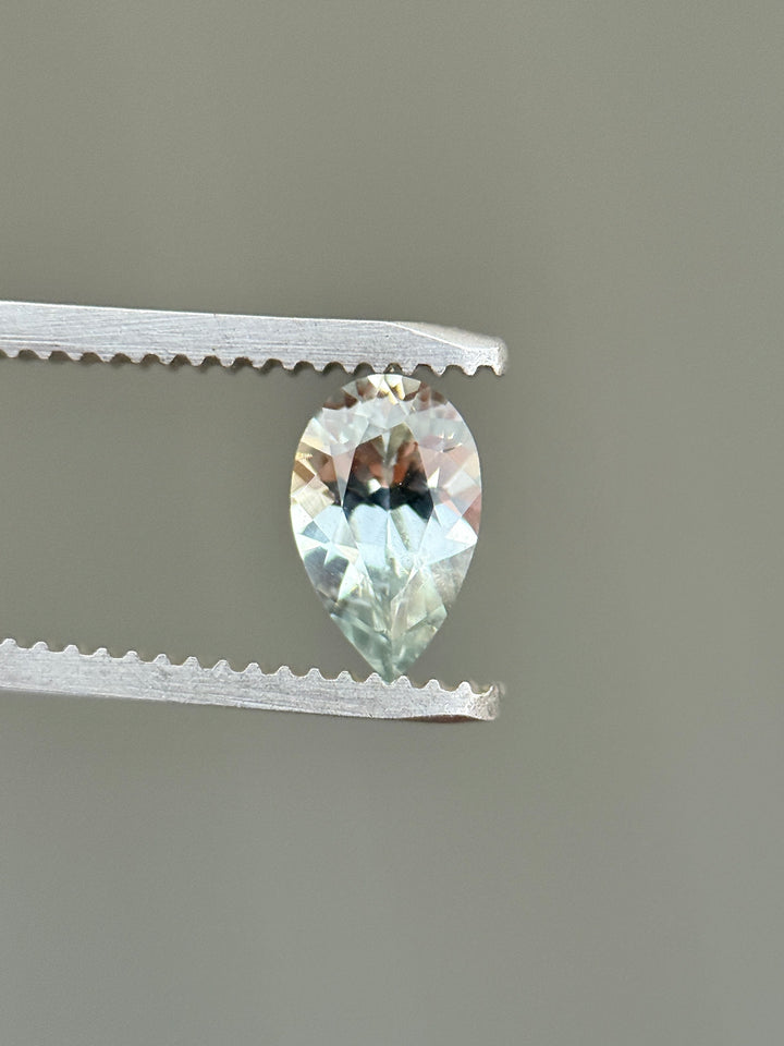 Side profile of a 1.13ct Montana sapphire held in gem tweezers, highlighting the precision pear mixed cut, pavilion depth, and subtle silver seafoam hue in an untreated natural sapphire.