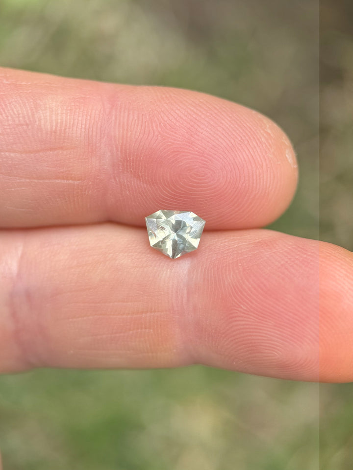 1.24ct parti sapphire with a custom shield brilliant cut, showcasing pastel green and yellow tones, balanced delicately on the fingertip with blurred grass in the background.