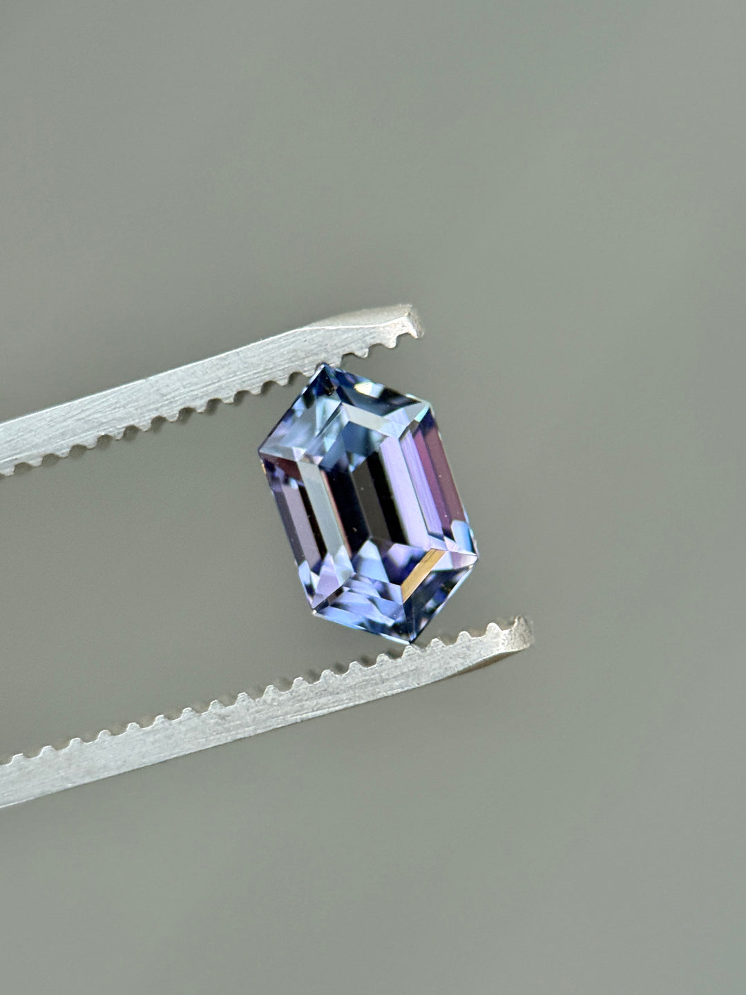Close-up of a 1.43-carat hexagon step cut tanzanite held in gem tweezers, showing strong multicolor mermaid tones—blue, green, pink, and purple—and untreated brilliance from Merelani Hills, Tanzania.