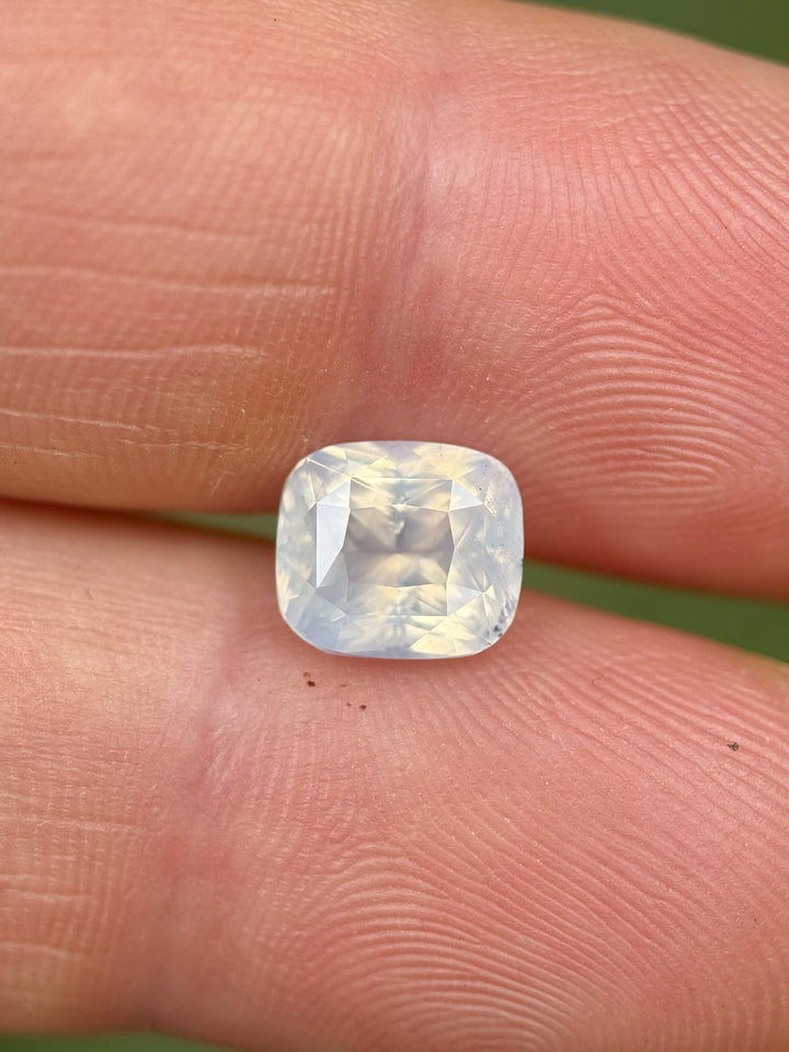 Opalescent 3.01 carat white sapphire placed on fingertip for size comparison, showcasing clarity, cut symmetry, and glowing white translucence, untreated cushion mixed cut sapphire from Ratnapura, Sri Lanka.