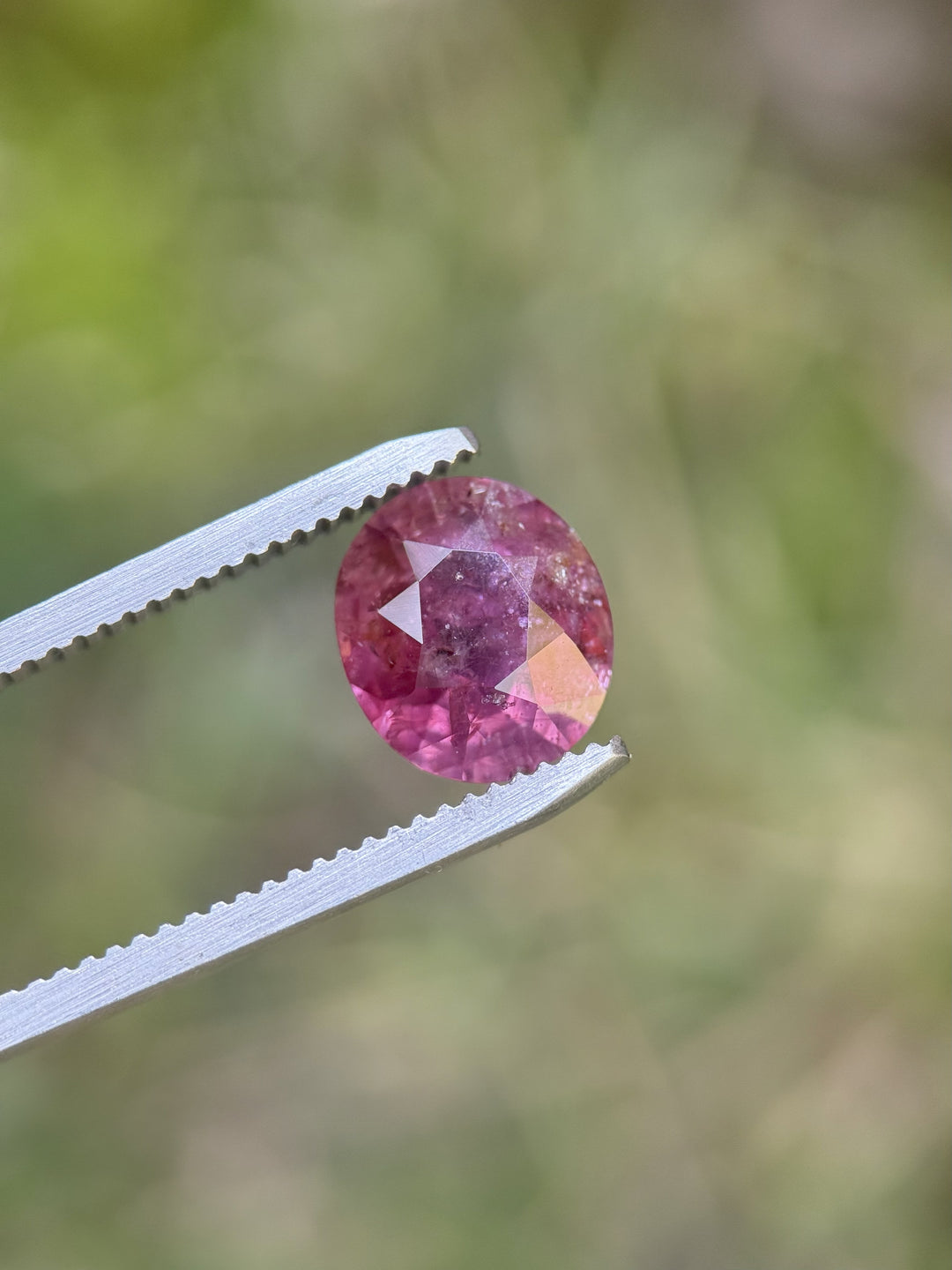 Custom oval cut 3.60 carat purplish red Tanzanian sapphire held in jeweler's tweezers with blurred green background