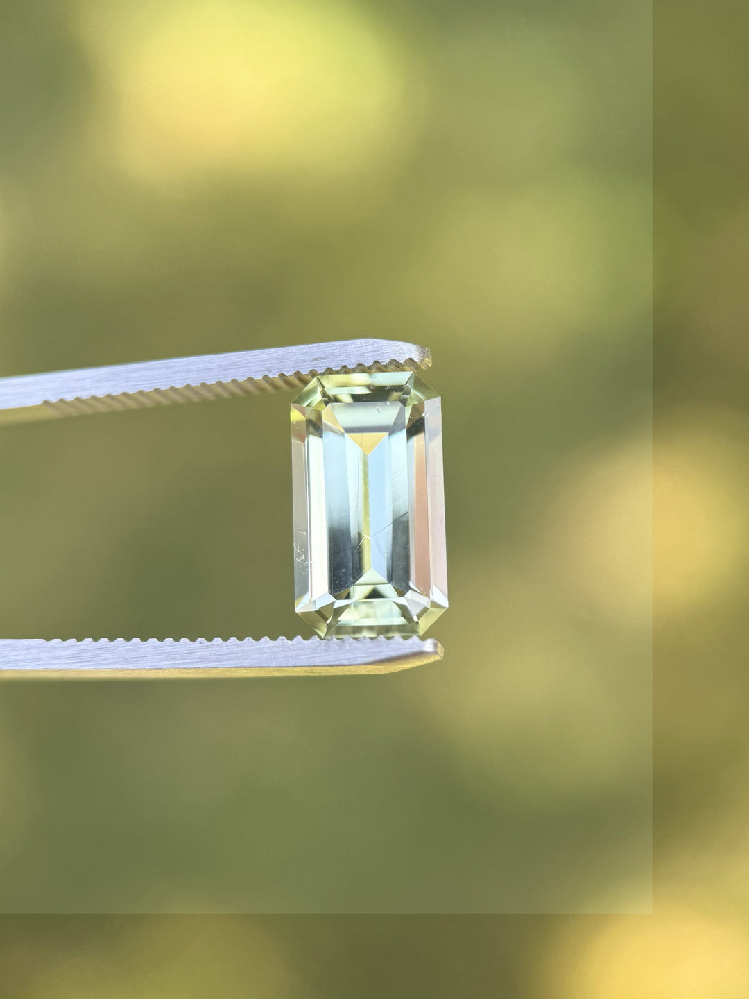 Mint green tourmaline weighing 4.51 carats held in gemstone tweezers against blurred natural background, showcasing sharp emerald cut geometry and exceptional clarity, no-heat gemstone from Congo, Africa.