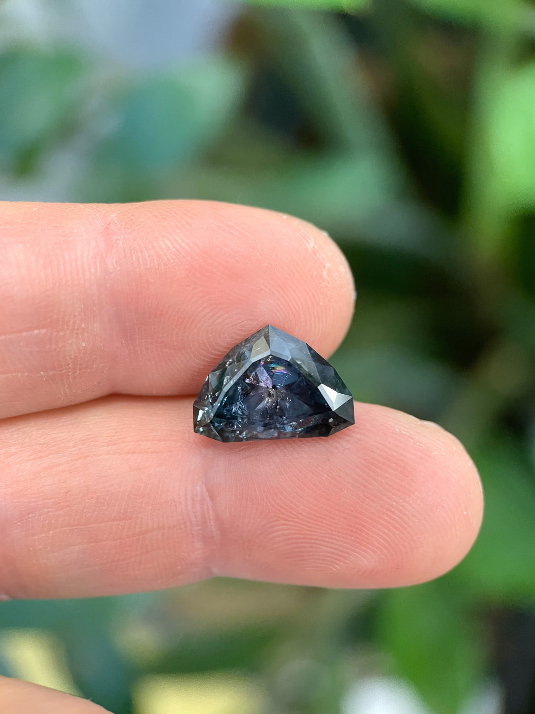 A 7.06-carat Montana sapphire with bluish-green tones shifting to gray-purple, displayed on fingertips. Modified shield brilliant cut.