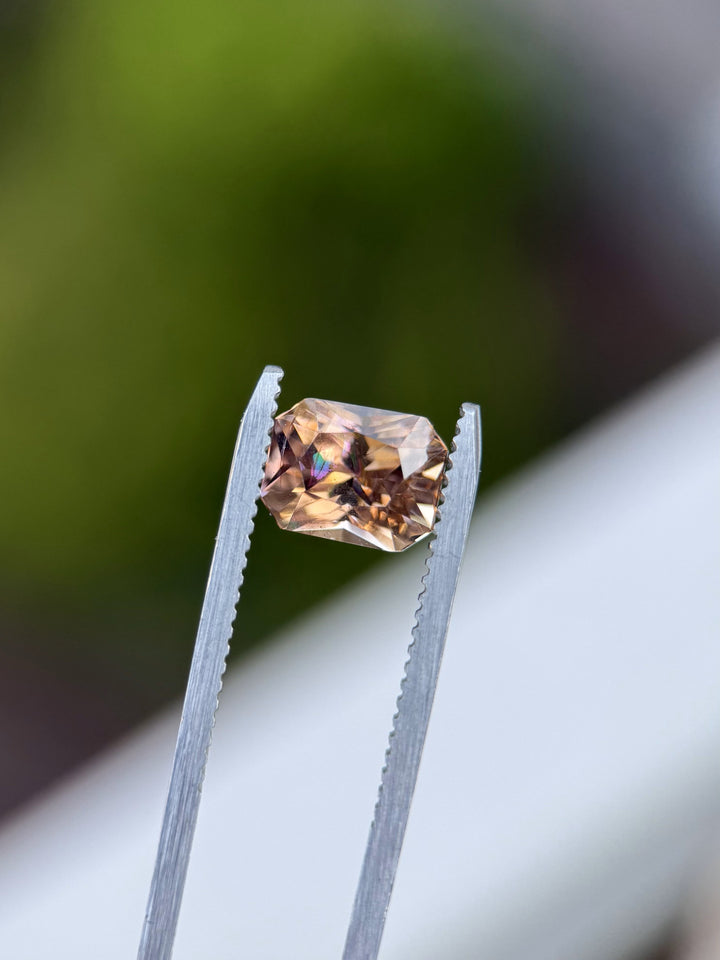 A 2.51 carat Tanzanite in a radiant cut held at an angle in tweezers, glowing with multicolor reflections of peach, purple, and champagne.