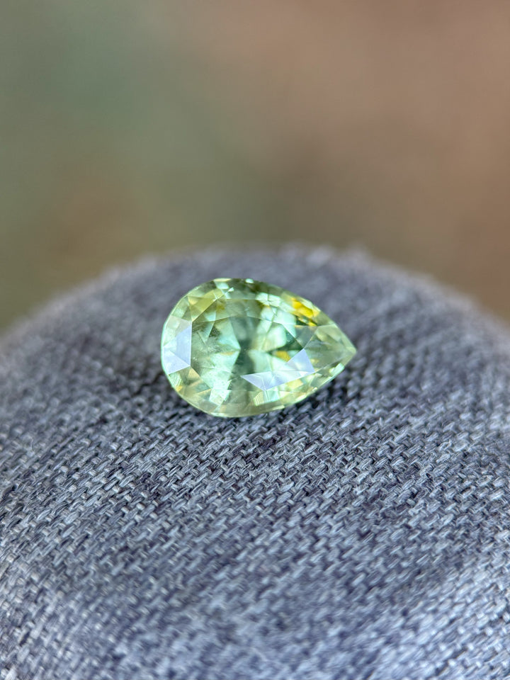 Close-up of a 3.04-carat sage green and peach sapphire in pear cut, placed on textured gray fabric with a muted brown background.