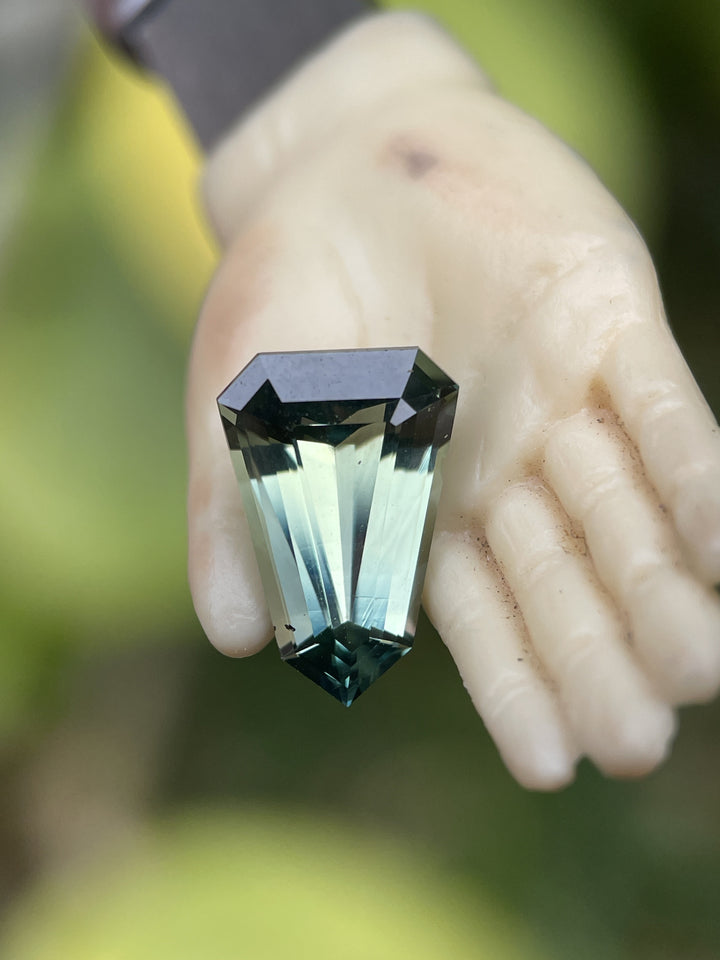 4.07 carat blue-green sapphire in a step cut shield shape shown in a small figurine’s hand, emphasizing the gemstone’s brilliance and unique color zoning.