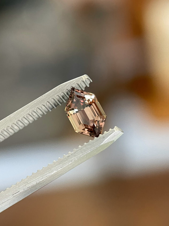 A precision-cut 1.22-carat Songea sapphire, delicately held in tweezers to display its hexagonal shape and captivating mix of brown, orange, and pink colors.