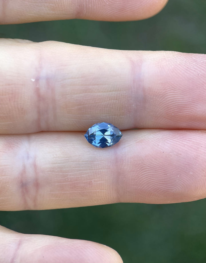 1.36 carat untreated sapphire in a custom geometric marquise cut showing a rich blue and periwinkle color, photographed against fingers for scale.