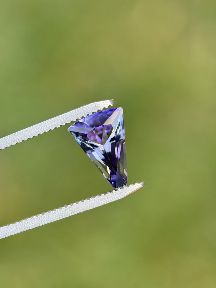 Custom fan cut Tanzanite gemstone with teal and purple hues held by gem tweezers against soft green background