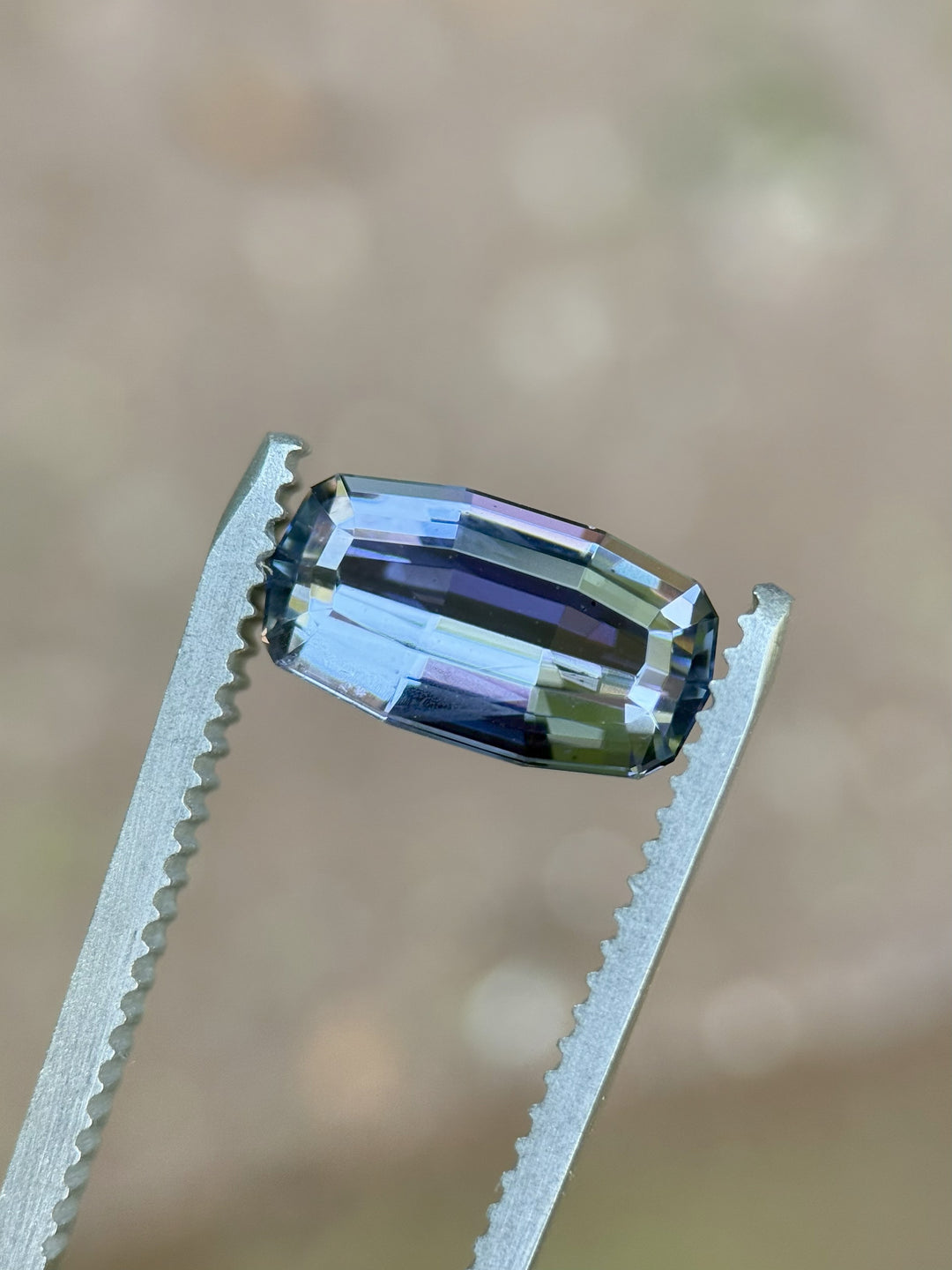 Modified emerald cut 2.63ct Tanzanite held in tweezers, displaying green, blue, and purple tones in natural lighting