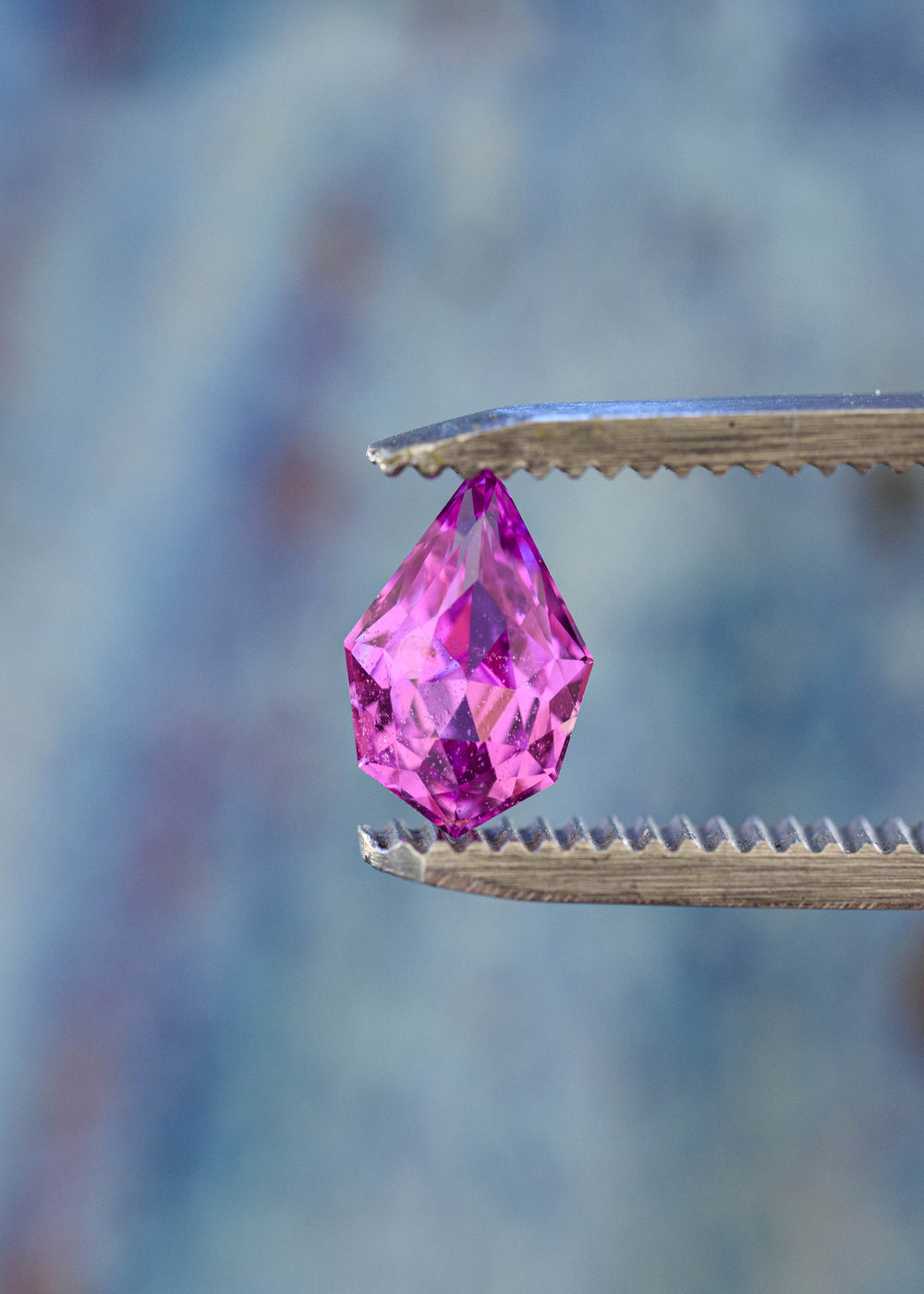 Untreated pink-purple Madagascar sapphire held upright in jeweler’s tweezers, highlighting its pointed geometric pear silhouette and angular precision faceting.