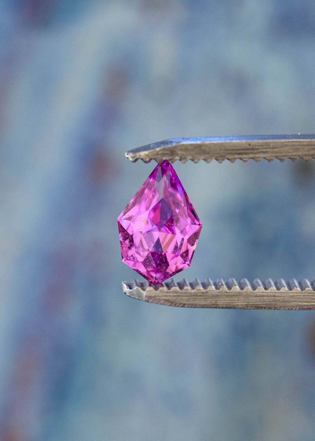 Untreated pink-purple Madagascar sapphire held upright in jeweler’s tweezers, highlighting its pointed geometric pear silhouette and angular precision faceting.