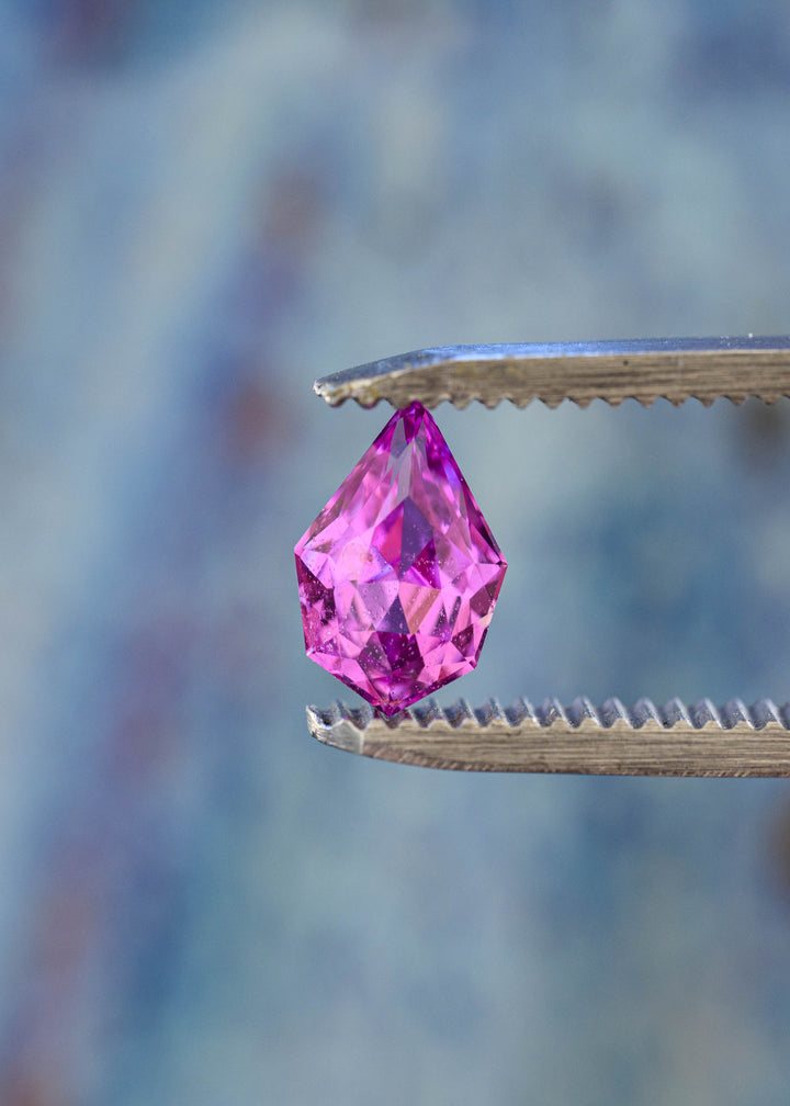 Untreated pink-purple Madagascar sapphire held upright in jeweler’s tweezers, highlighting its pointed geometric pear silhouette and angular precision faceting.