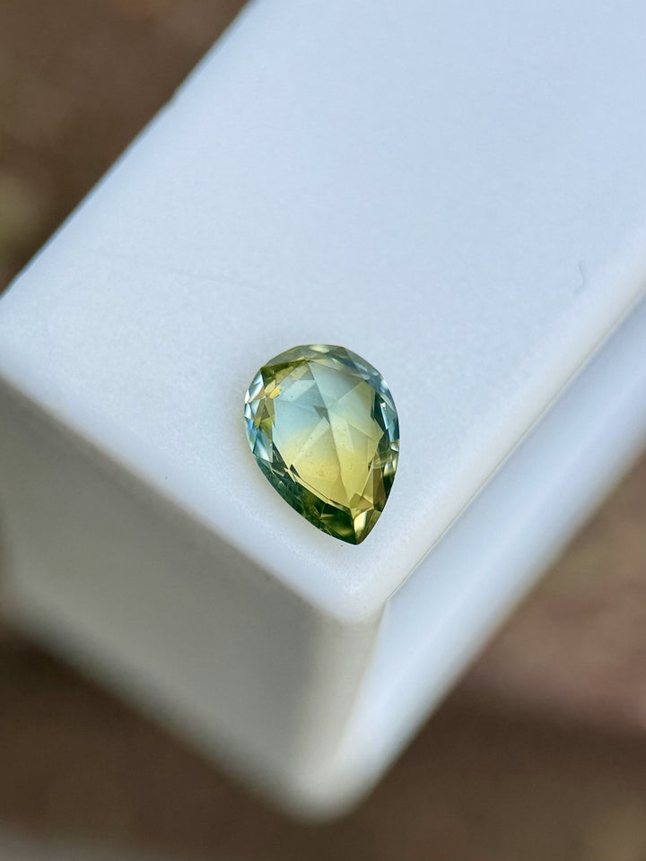 1.07-carat parti sapphire resting on a white display box showing vibrant teal and yellow hues.