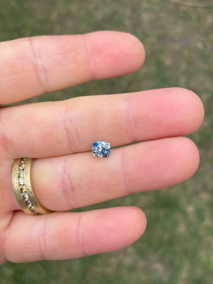 "A 1.78-carat Madagascar Parti Sapphire resting on a finger, showcasing its modified brilliant cushion cut and striking combination of colors, including yellow, colorless, and various shades of blue.