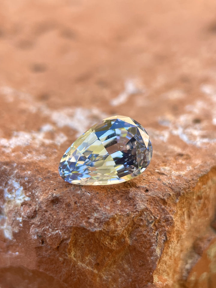 A stunning 2.39-carat custom mix brilliant teardrop sapphire placed on a rustic stone, showcasing brilliant cornflower and powder blue hues. This gemstone measures 9.60mm in length, 6.54mm in width, and 4.80mm in height, and is untreated, sourced from Sri Lanka.