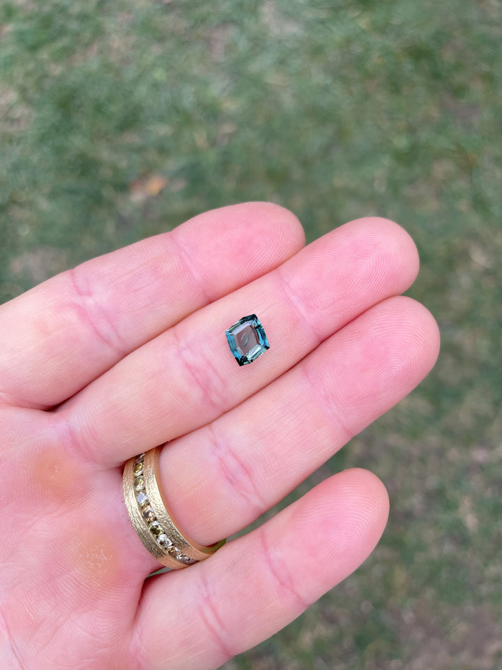 Close-up image of a stunning teal and blue 1.68-carat no heat sapphire, cut in a unique custom geometric rhombus portrait style, held on a finger against a natural outdoor background. The gemstone showcases brilliant color saturation and fine craftsmanship.