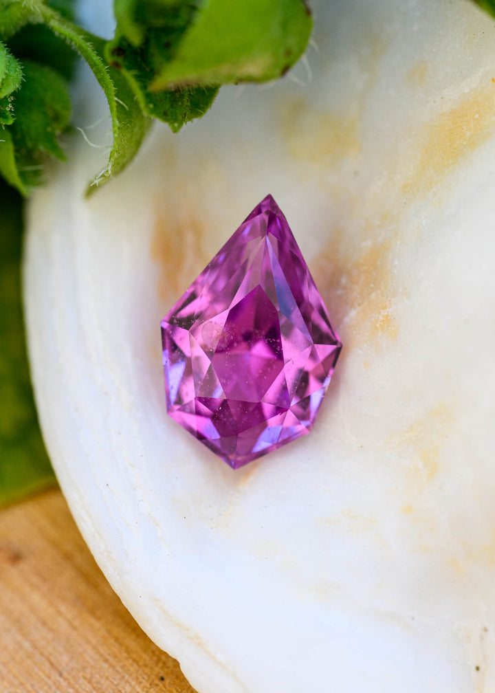 Vibrant pink-purple 1.27 carat sapphire with a precision geometric pear cut resting against a white flower, showing saturated fuchsia and violet facets.
