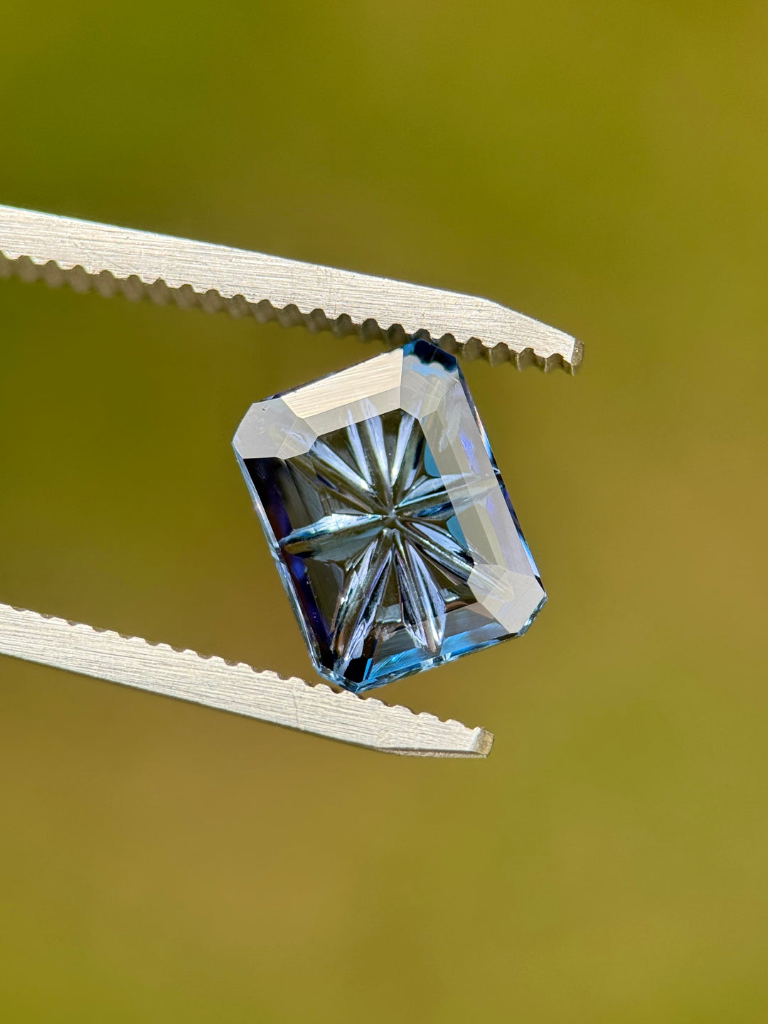 Untreated 2.99ct Tanzanite showing violet, teal, and blue tones in an octagonal concave portrait cut held in gem tweezers, highlighting the detailed starburst concave faceting.