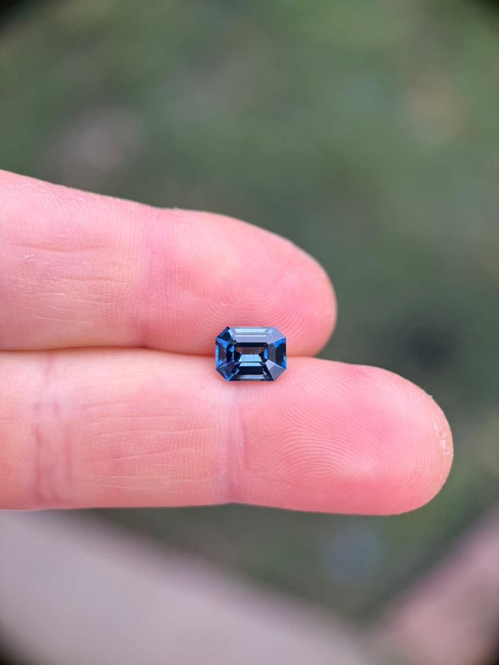 Teal indigo sapphire emerald cut on fingertip showing size scale, clean faceting, and vivid color from Madagascar.