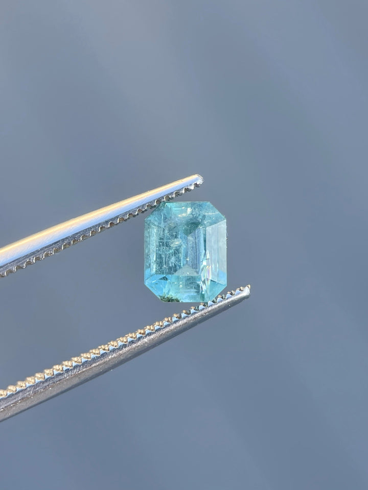 A 1.17-carat emerald gemstone, emerald cut, with vibrant green-blue hues held in precision tweezers.