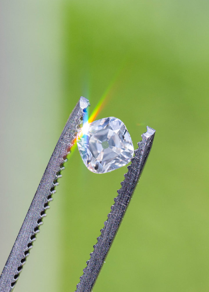 White antique-style cushion diamond held in jeweler's tweezers with sunlight creating colorful dispersion and highlighting its symmetrical Old Mine inspired faceting