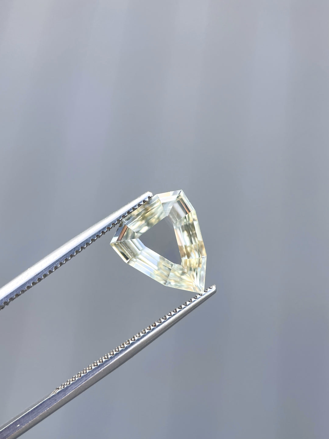 A 2.47-carat yellow-green sapphire with a modified geometric step cut, held by tweezers, showcasing its unique portrait shield shape. The gemstone's clean facets reflect light beautifully. The sapphire, sourced from Sri Lanka, measures 11.68mm by 9.62mm by 2.63mm and is untreated.