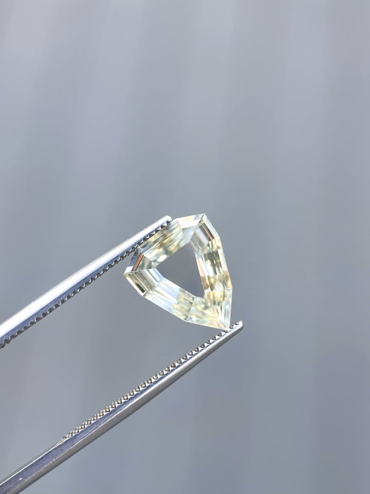 A 2.47-carat yellow-green sapphire with a modified geometric step cut, held by tweezers, showcasing its unique portrait shield shape. The gemstone's clean facets reflect light beautifully. The sapphire, sourced from Sri Lanka, measures 11.68mm by 9.62mm by 2.63mm and is untreated.