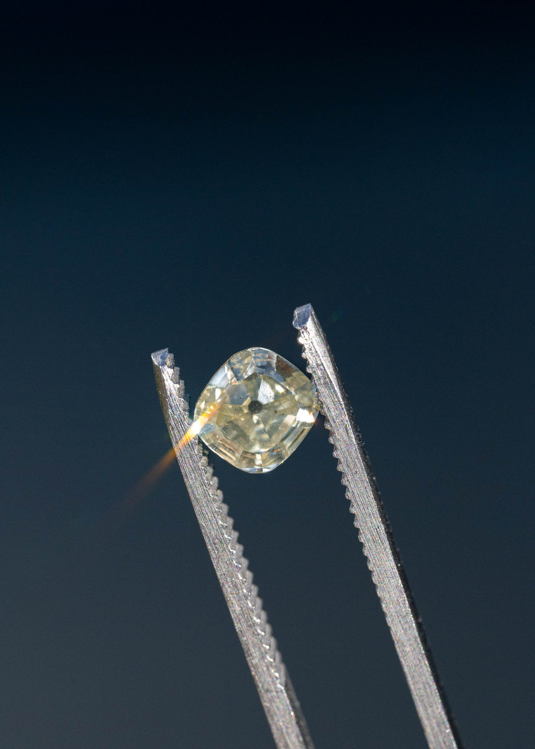 Yellow antique-style cushion diamond held in jeweler's tweezers against a dark background, showcasing bold flashes of light and its Old Mine inspired faceting.