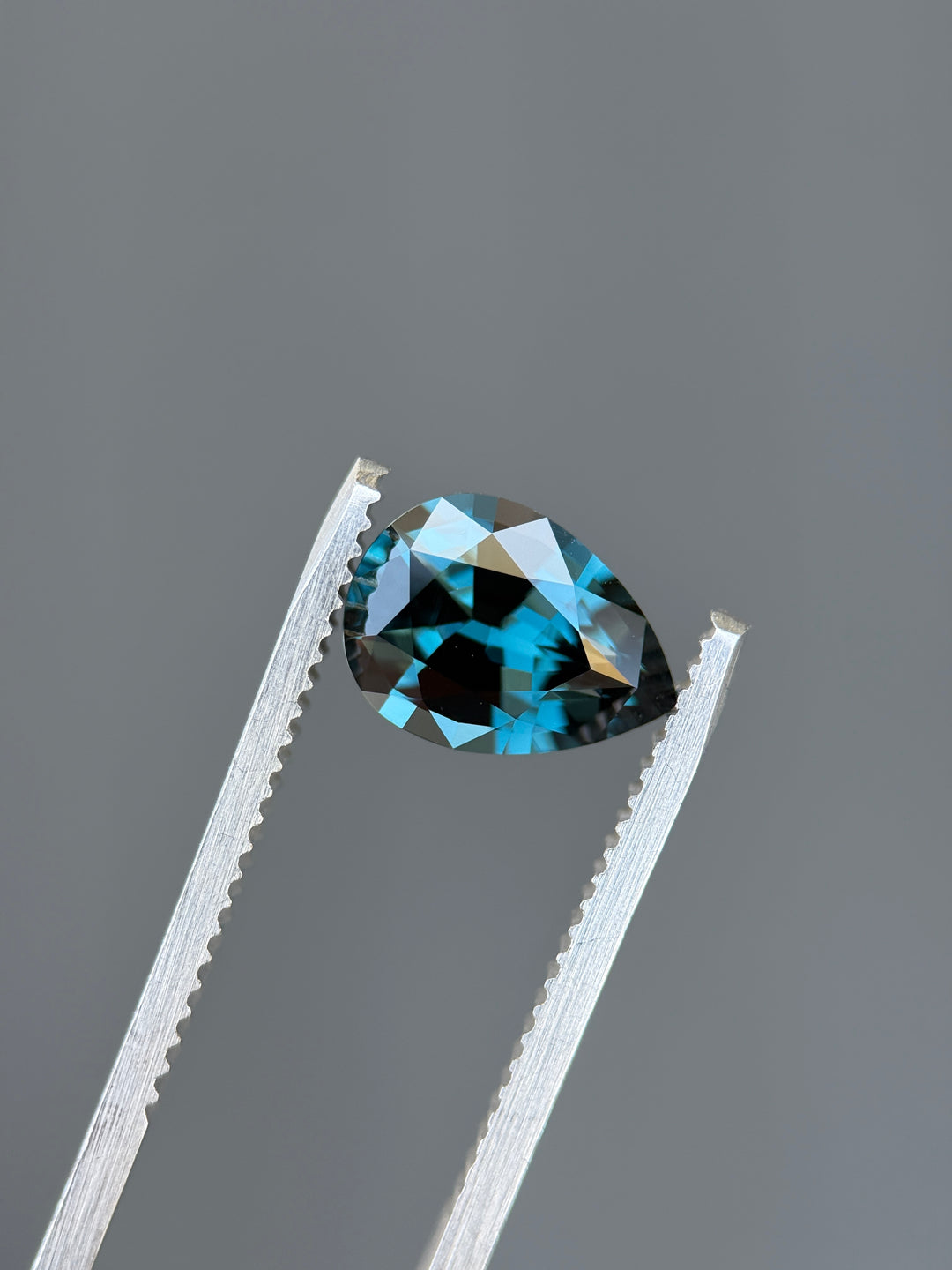 A faceted pear-shaped light blue gemstone held between silver metal tweezers against a smooth gray background.