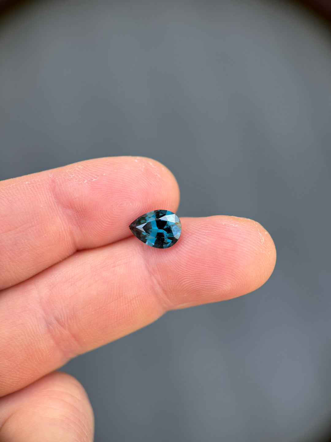 A small, faceted pear-cut gemstone with a vibrant blue and black mottled pattern, held between two fingers to show its size against a blurred gray background.