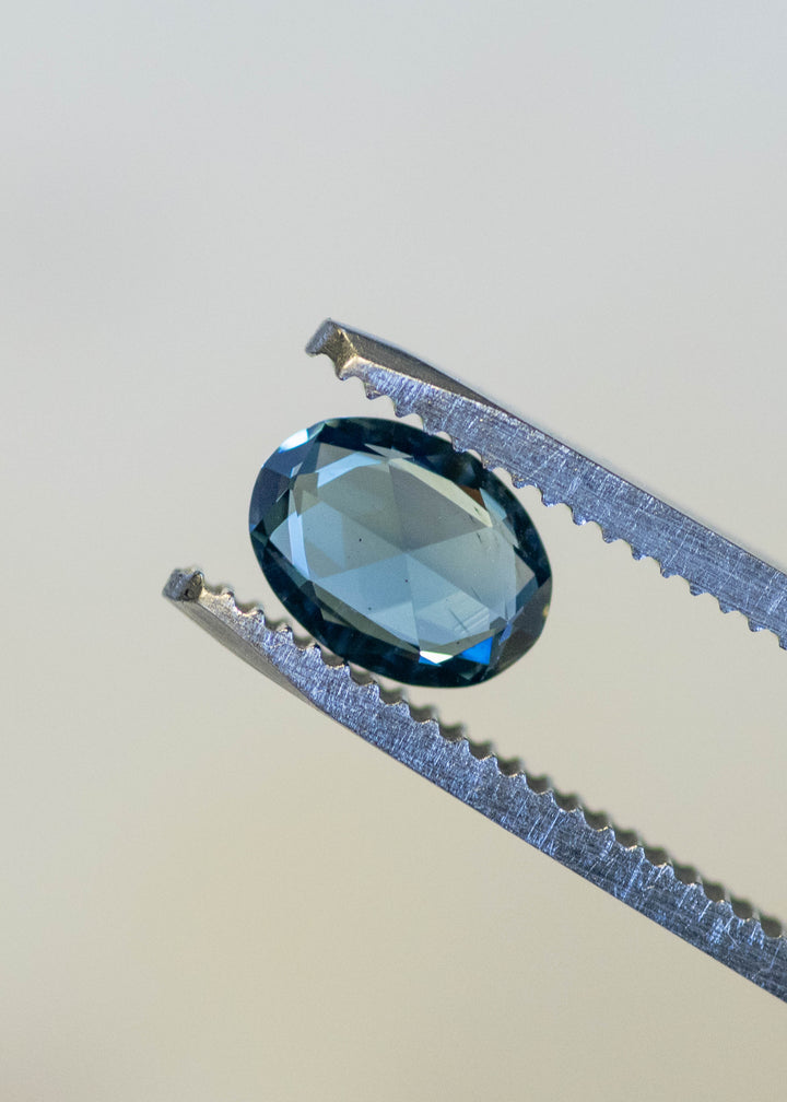 Close-up of a 1.37ct Madagascar sapphire resting on fingertips, revealing blue-green body color, rose-cut symmetry, and subtle teal flashes.