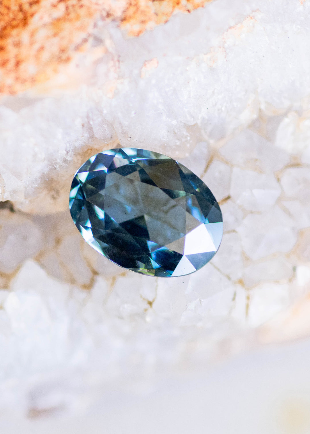 1.37 carat blue teal green sapphire nestled among soft green botanical buds, displaying an oval rose cut with geometric facets and shifting ocean-blue flashes.