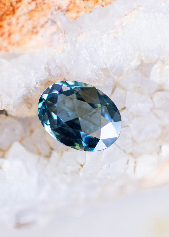 1.37 carat blue teal green sapphire nestled among soft green botanical buds, displaying an oval rose cut with geometric facets and shifting ocean-blue flashes.