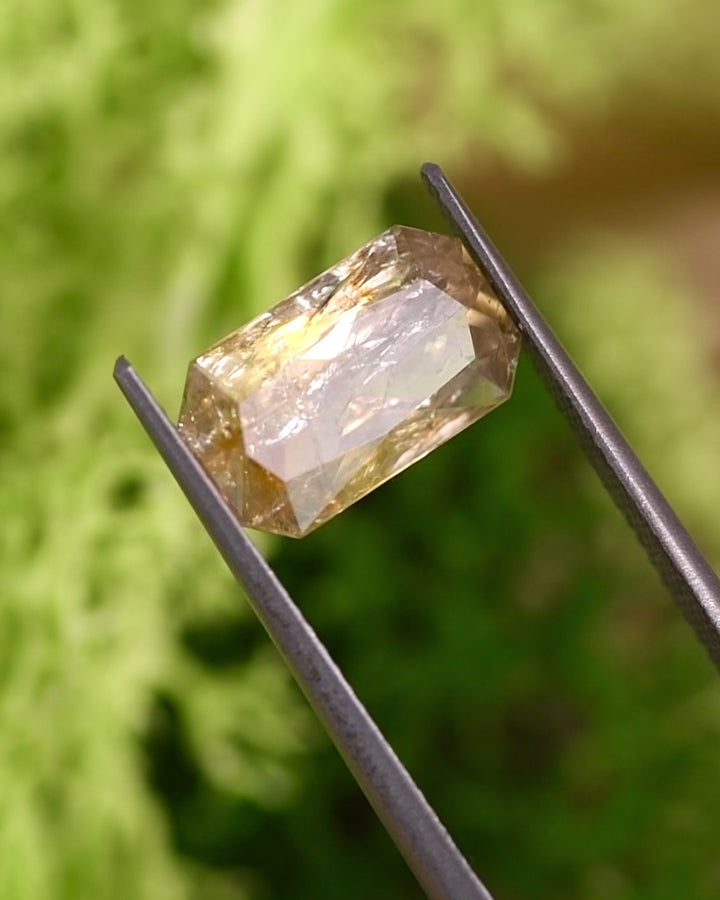 Video of 3.50CT yellow and orange sapphire turning in tweezers under natural sunlight, showing color shifts and sparkle through its custom modified radiant cut, untreated gemstone from Umba River Valley, Tanzania.