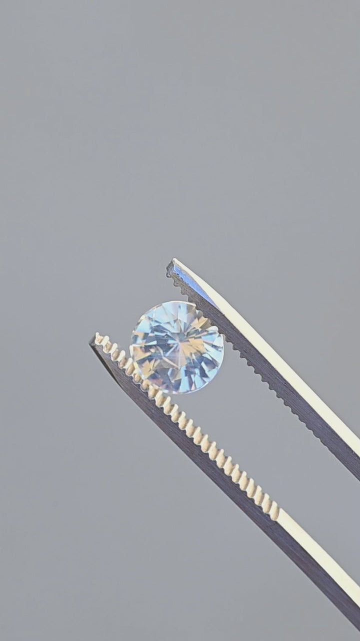Natural round white opalescent sapphire rotating in gem tweezers under natural lighting showing off the opalescent glow.