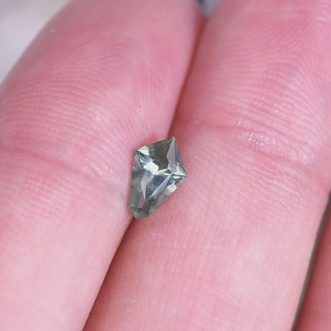 0.57 carat teal green Montana sapphire kite step cut placed on fingertip for scale, highlighting geometric shape, color zoning, and untreated clarity