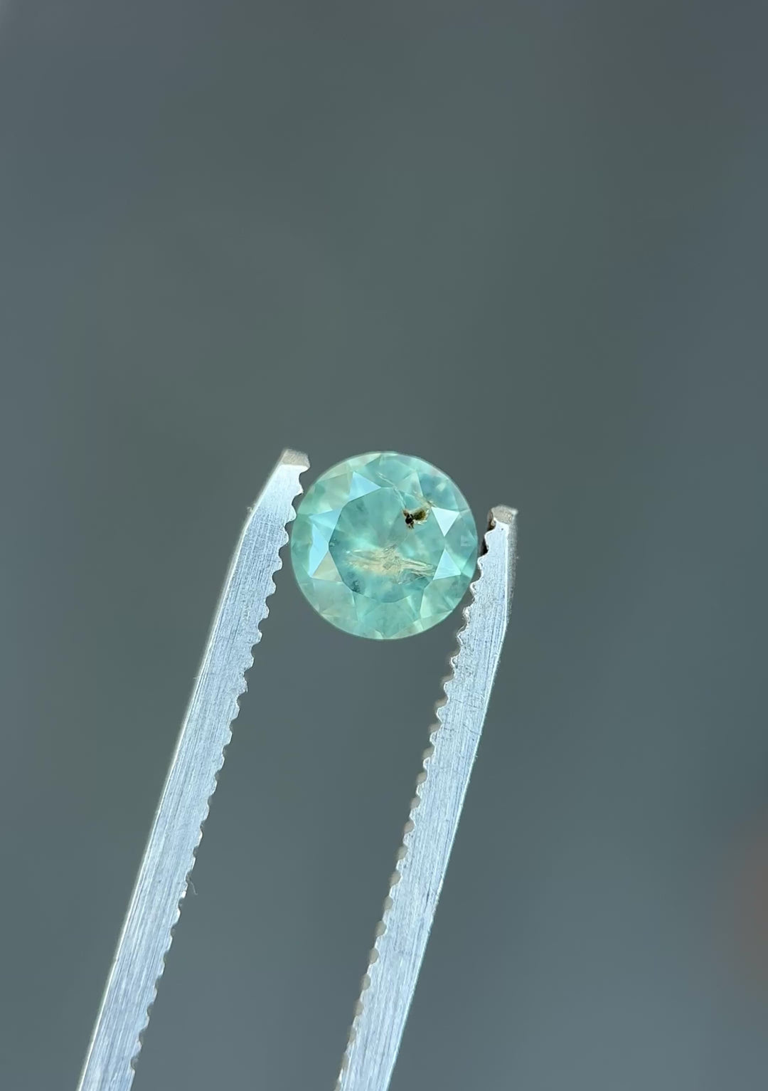 Natural Montana sapphire turning in tweezers, showcasing powder teal color, gentle opalescence, and symmetrical round faceting.
