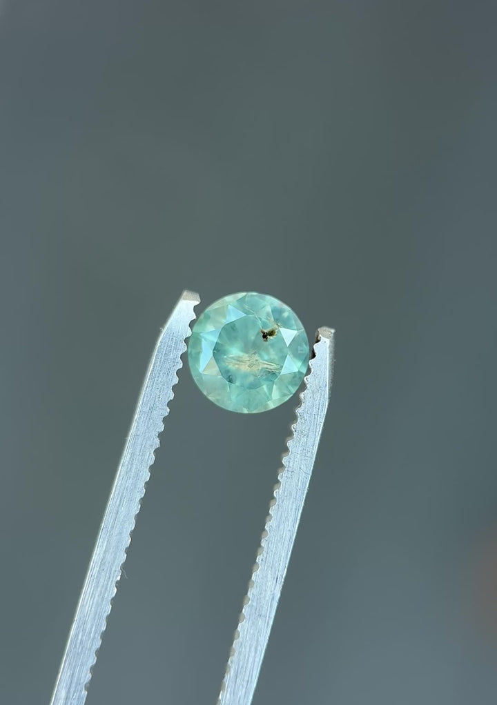Natural Montana sapphire turning in tweezers, showcasing powder teal color, gentle opalescence, and symmetrical round faceting.